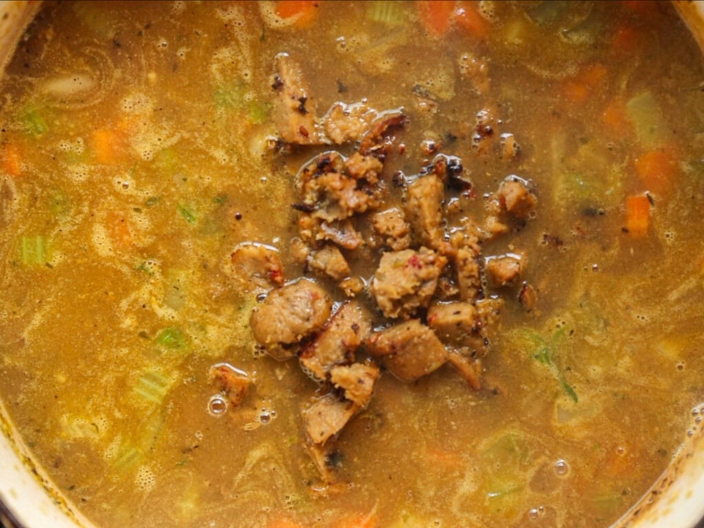A close-up of a hearty soup with chopped vegetables and pieces of cooked vegan sausage in a thick, brownish broth. The meat is piled in the center, surrounded by visible celery and carrots.